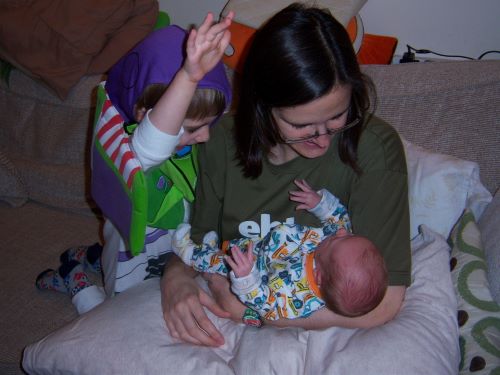 a white woman with brown hair and glasses sits on a couch and smiles down at a baby in her arms with a small child at her side